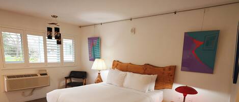 A bedroom with a wooden bed, white bedding, a nightstand with a lamp, a radiator, and a window with blinds.