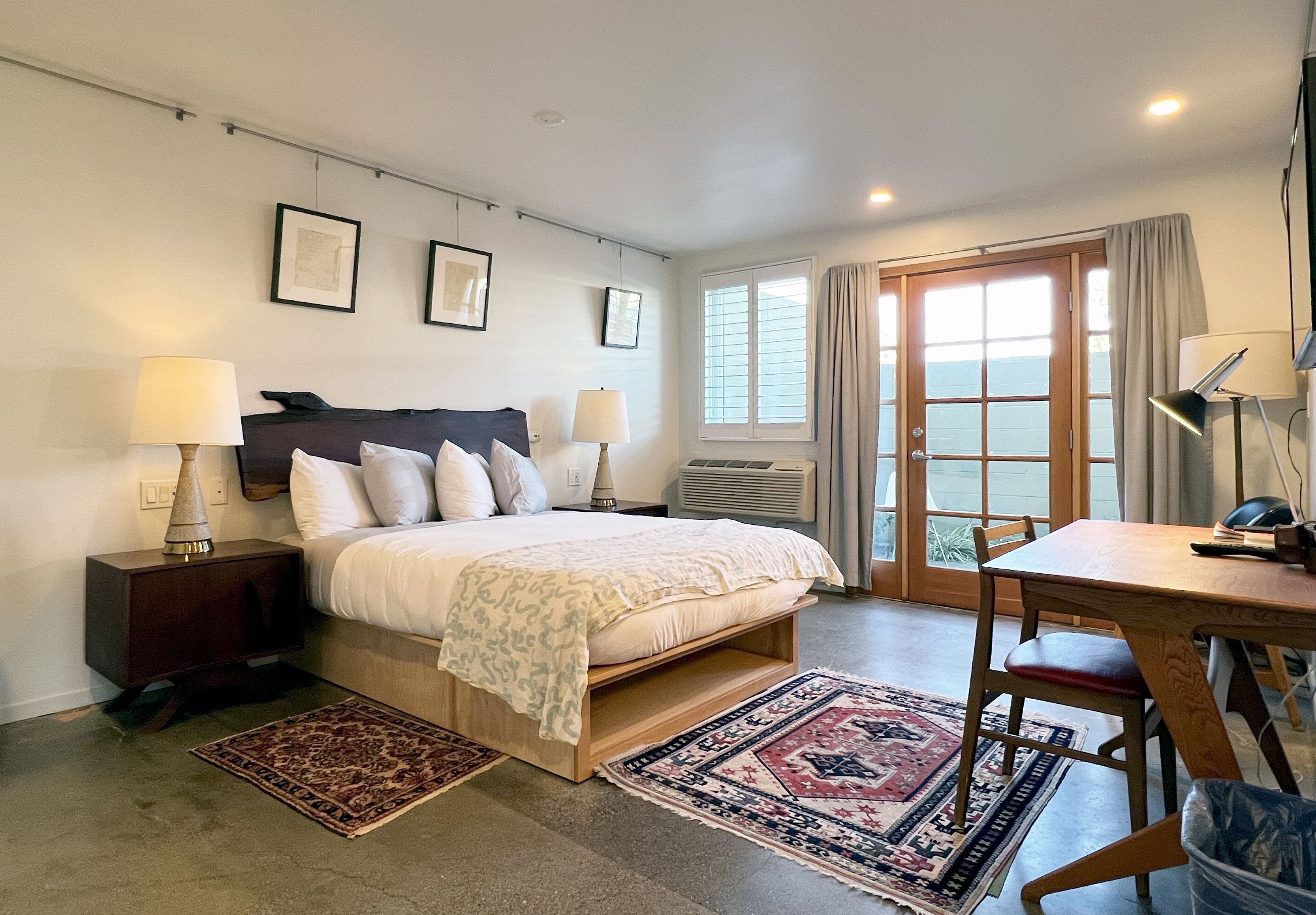 Un dormitorio con cama, mesitas de noche, escritorio y silla. Se ve una ventana con cortinas y vistas al exterior.