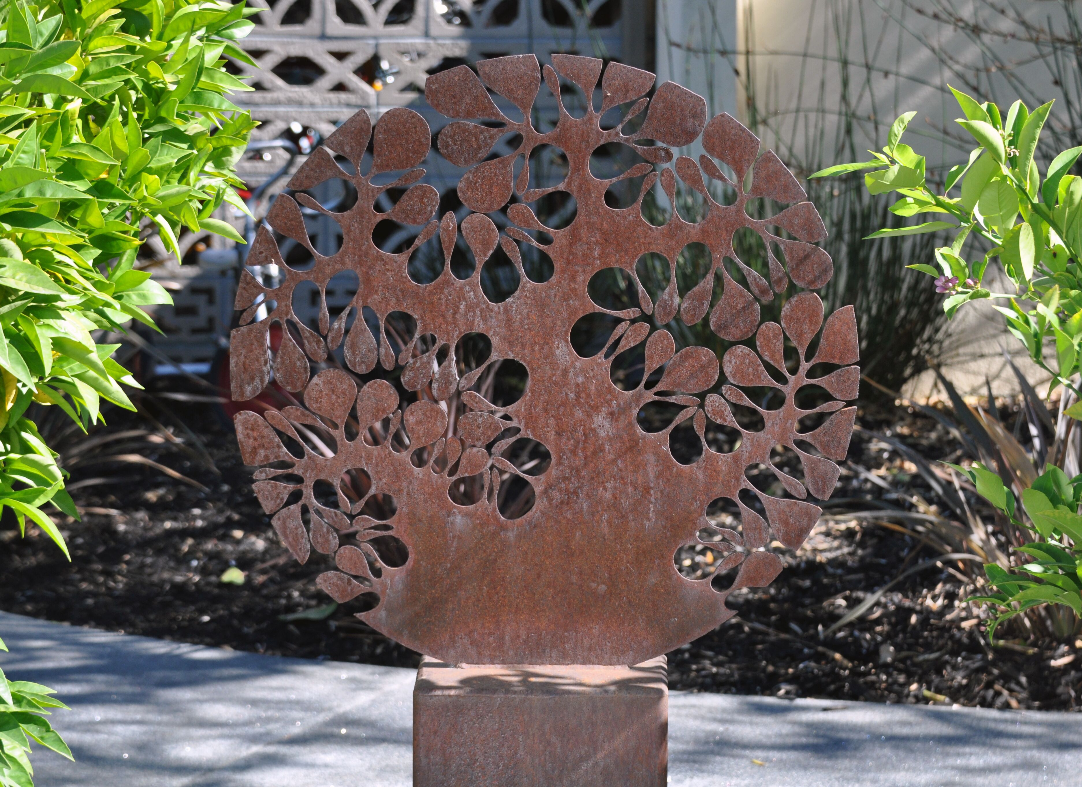 A metal sculpture with leaf-like cutouts, set on a concrete base, surrounded by greenery.