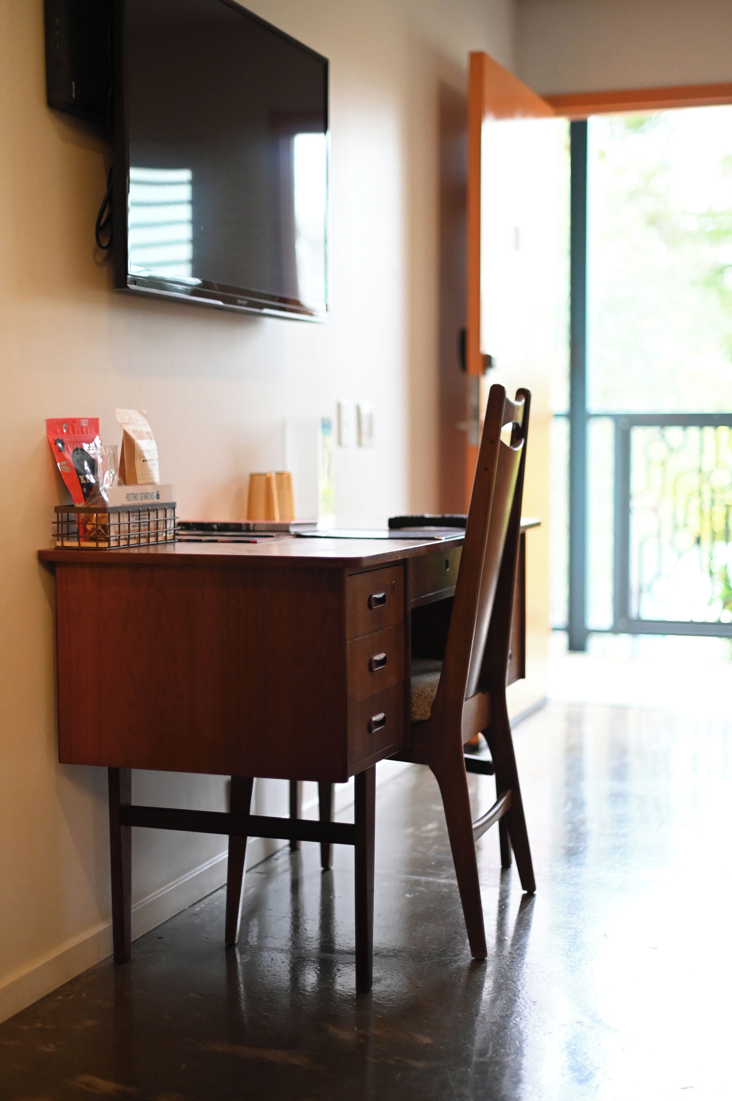 A wooden desk with a chair, a flat-screen TV mounted on the wall, and a window with a view of greenery.