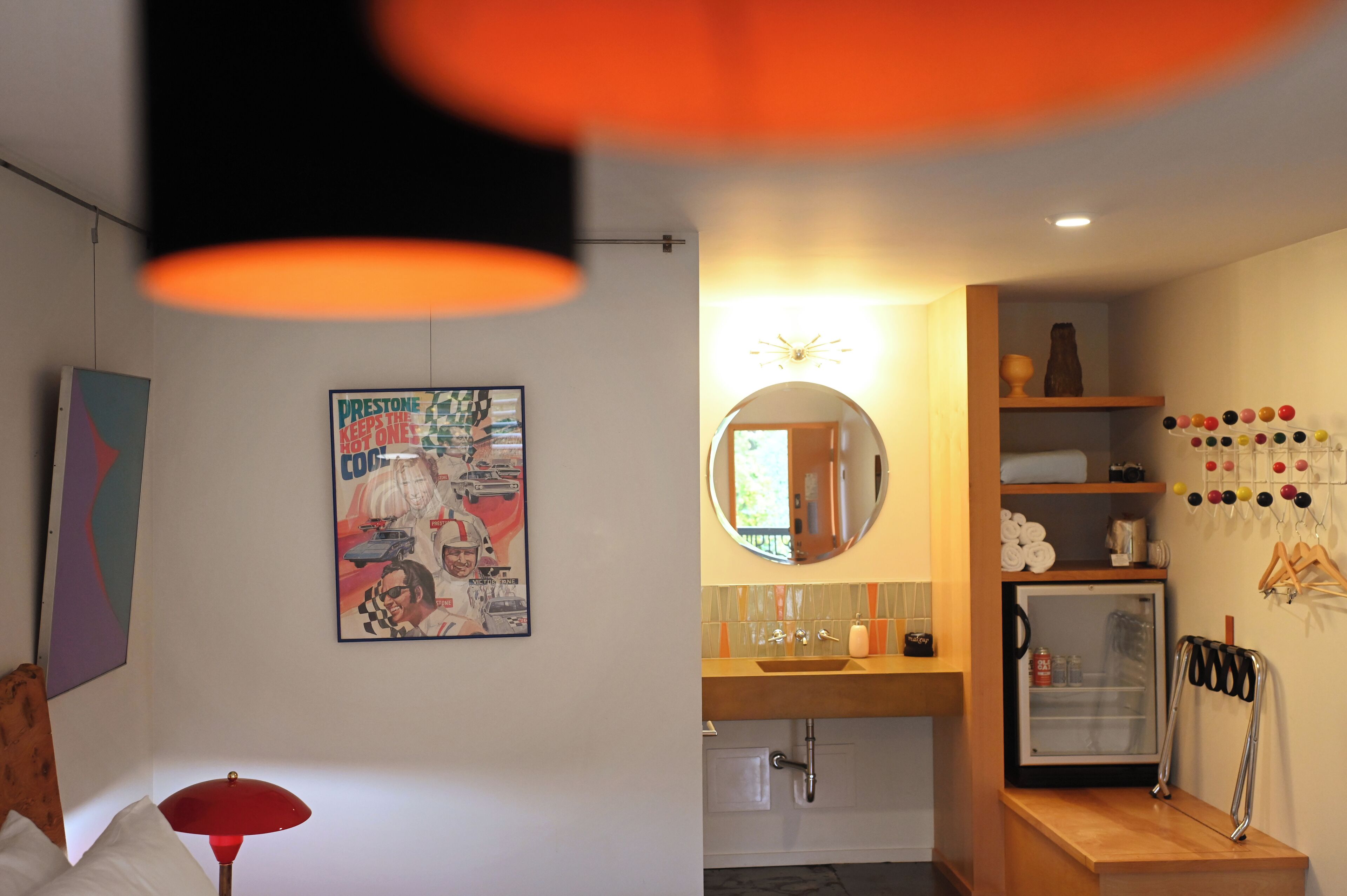 A modern bathroom with a round mirror, a sink, and a small refrigerator. There is a colorful poster on the wall and a lamp on the floor.