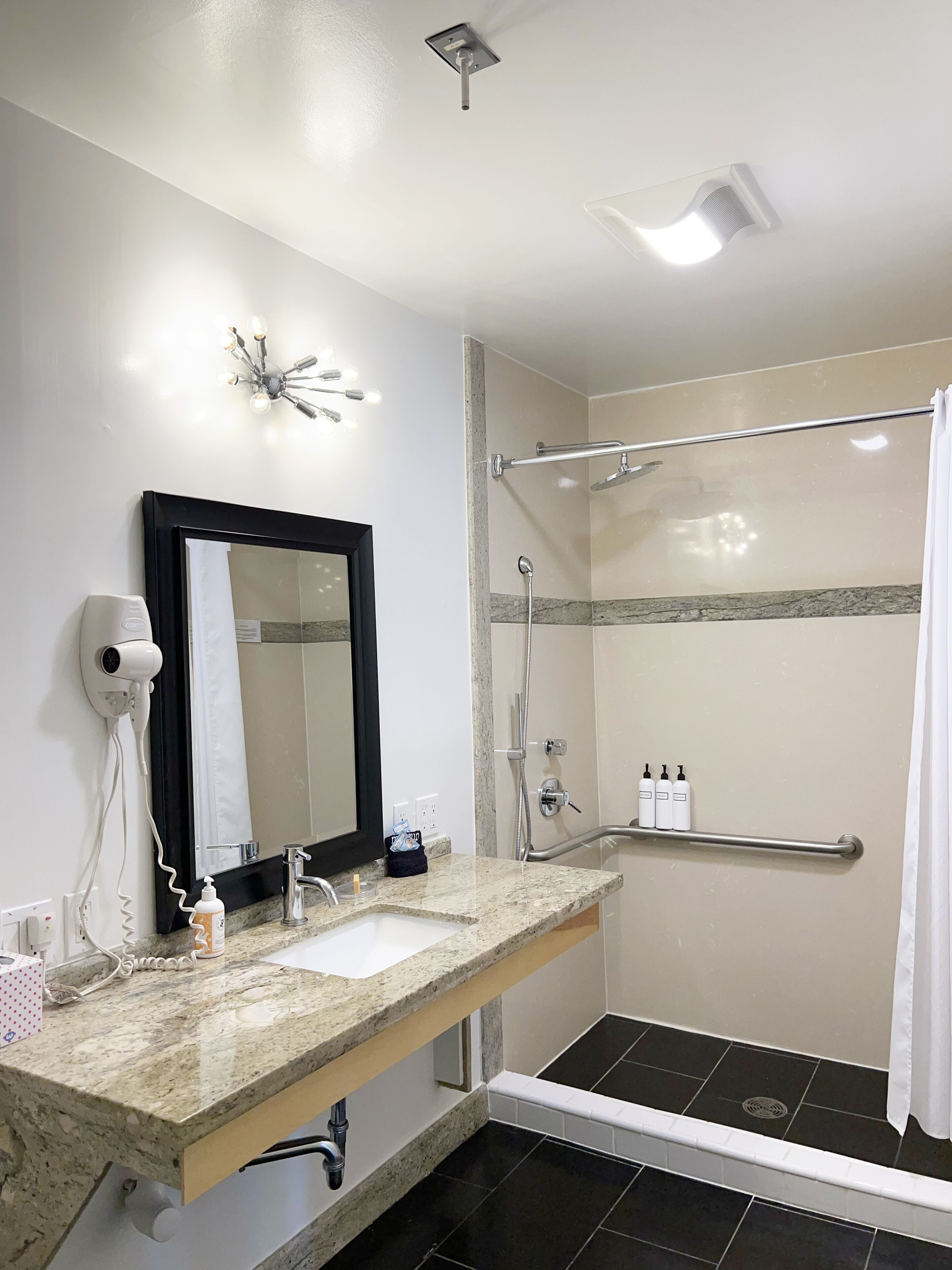 A bathroom with a sink, mirror, hairdryer, and shower with a grab bar.