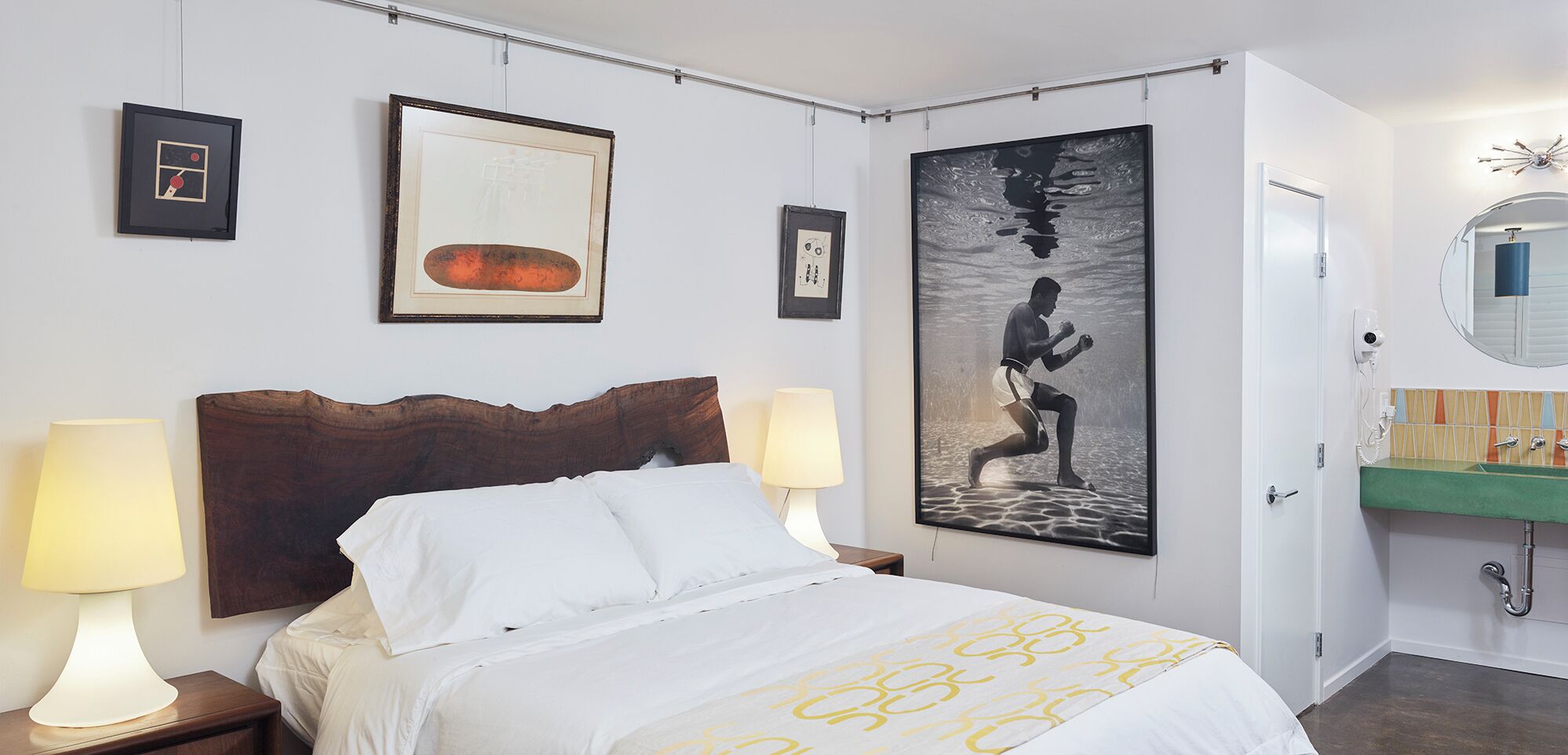 A hotel room with a bed, two bedside lamps, a large framed artwork, a bathroom with a mirror and sink, and a door.