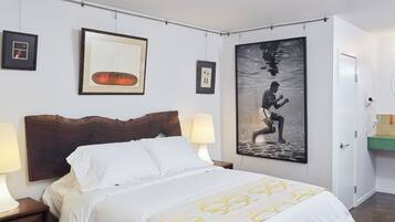 A hotel room with a bed, two bedside lamps, a large framed artwork, a bathroom with a mirror and sink, and a door.