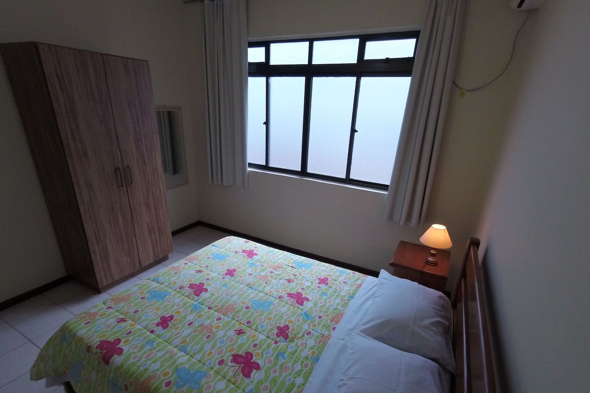 A bedroom with a bed, a wardrobe, a window, and a nightstand with a lamp.
