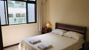 A hotel room with a bed, a nightstand with a lamp, an air conditioning unit, and a window with curtains. - Regina Del Mare Residence (Florianópolis)