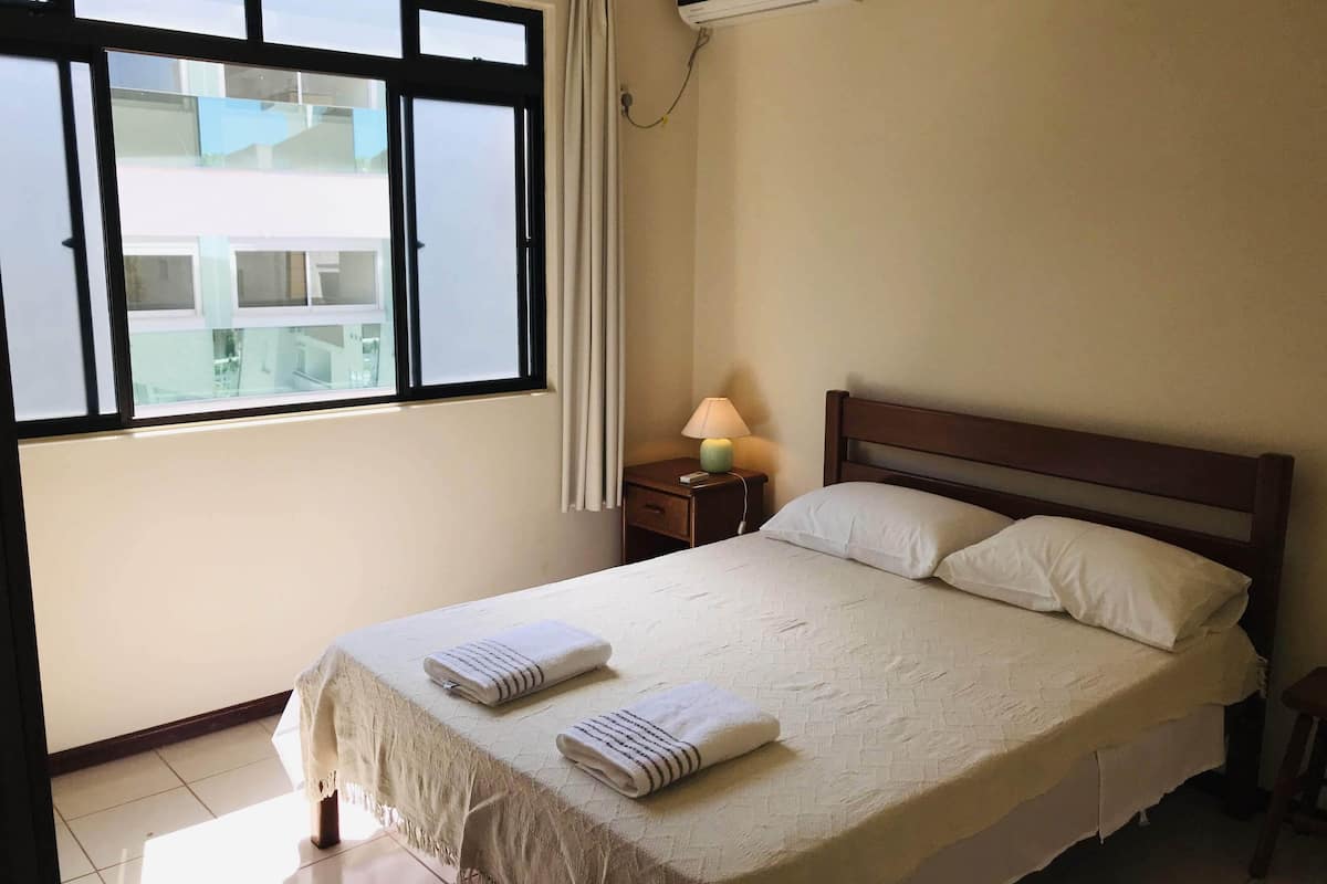 A hotel room with a bed, a nightstand with a lamp, an air conditioning unit, and a window with curtains.