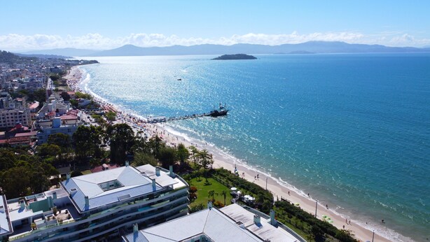 A coastal cityscape with a beach, a pier, and modern buildings. - Regina Del Mare Residence (Florianópolis)