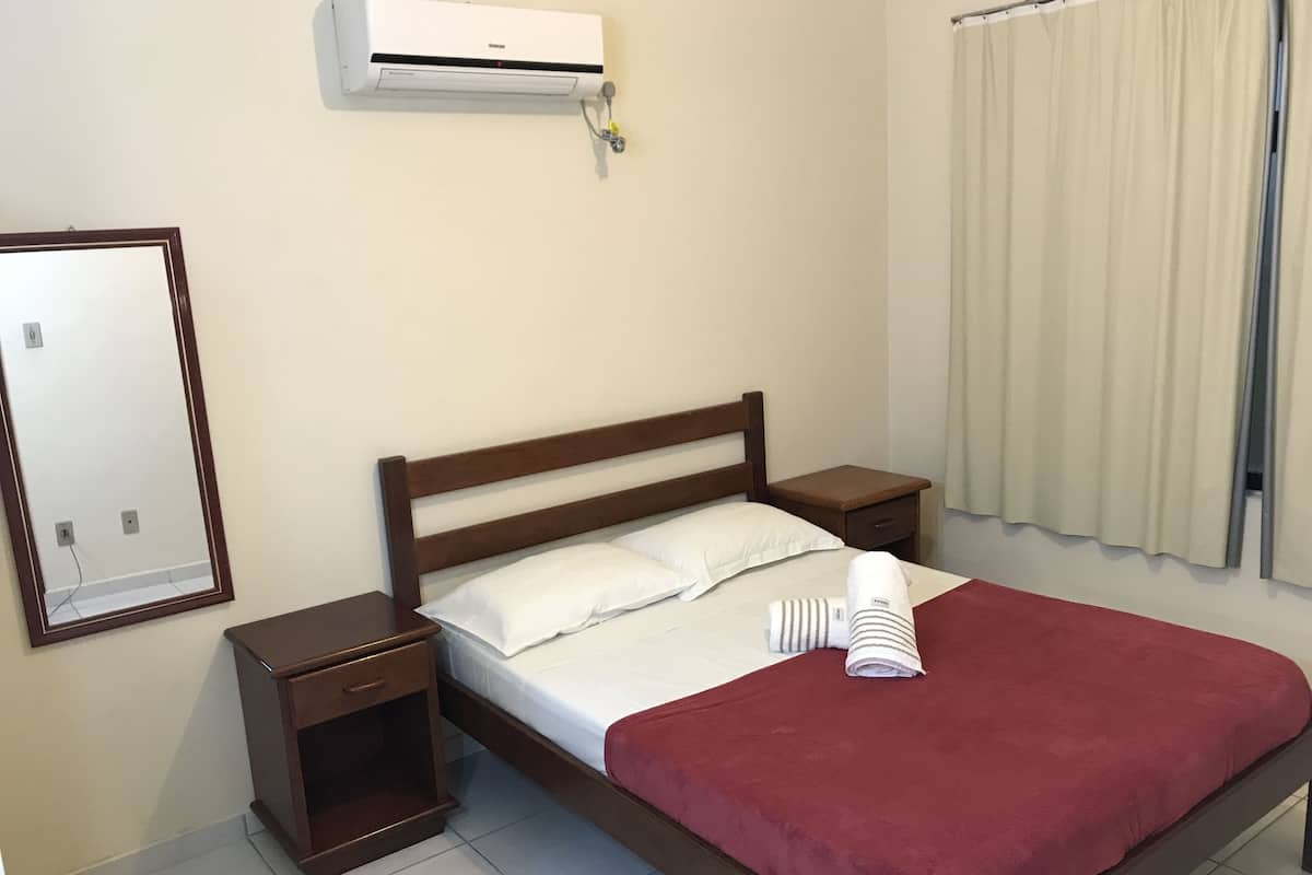 A hotel room with a bed, a nightstand, a drawer, a mirror, an air conditioning unit, and a window with curtains.