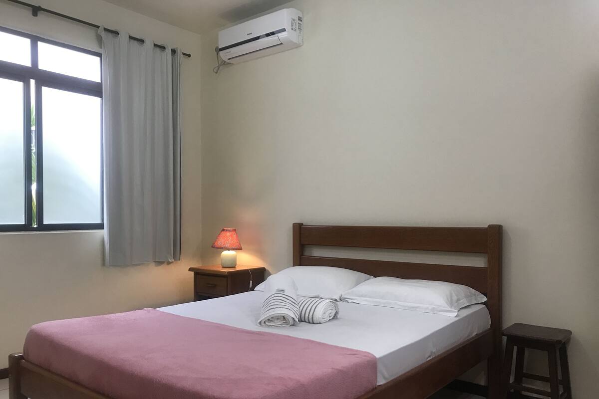 A neatly made bed with a pink blanket, a bedside lamp, and a small stool.