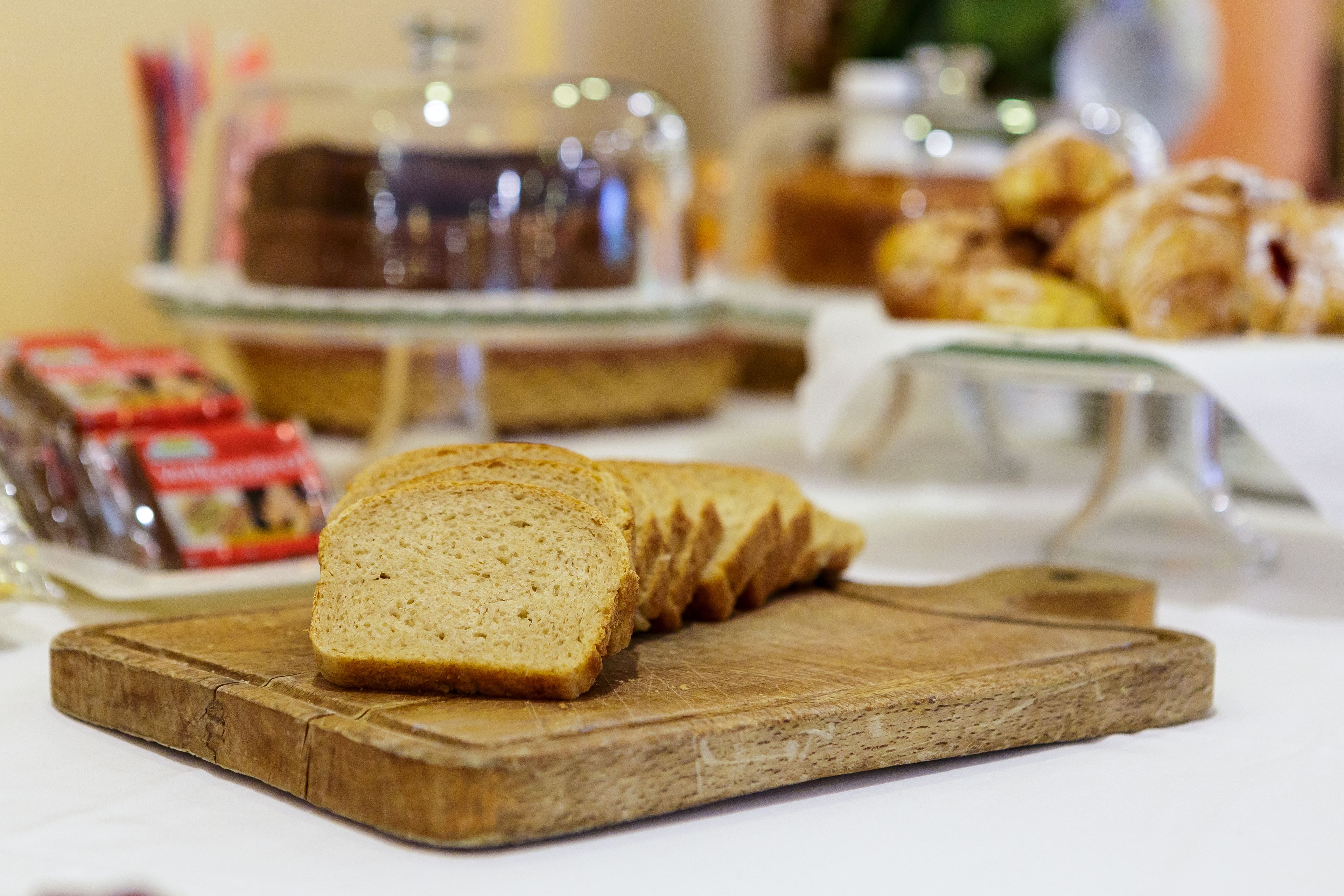 Slices of bread on a wooden board, with a glass container and pastries in the background.