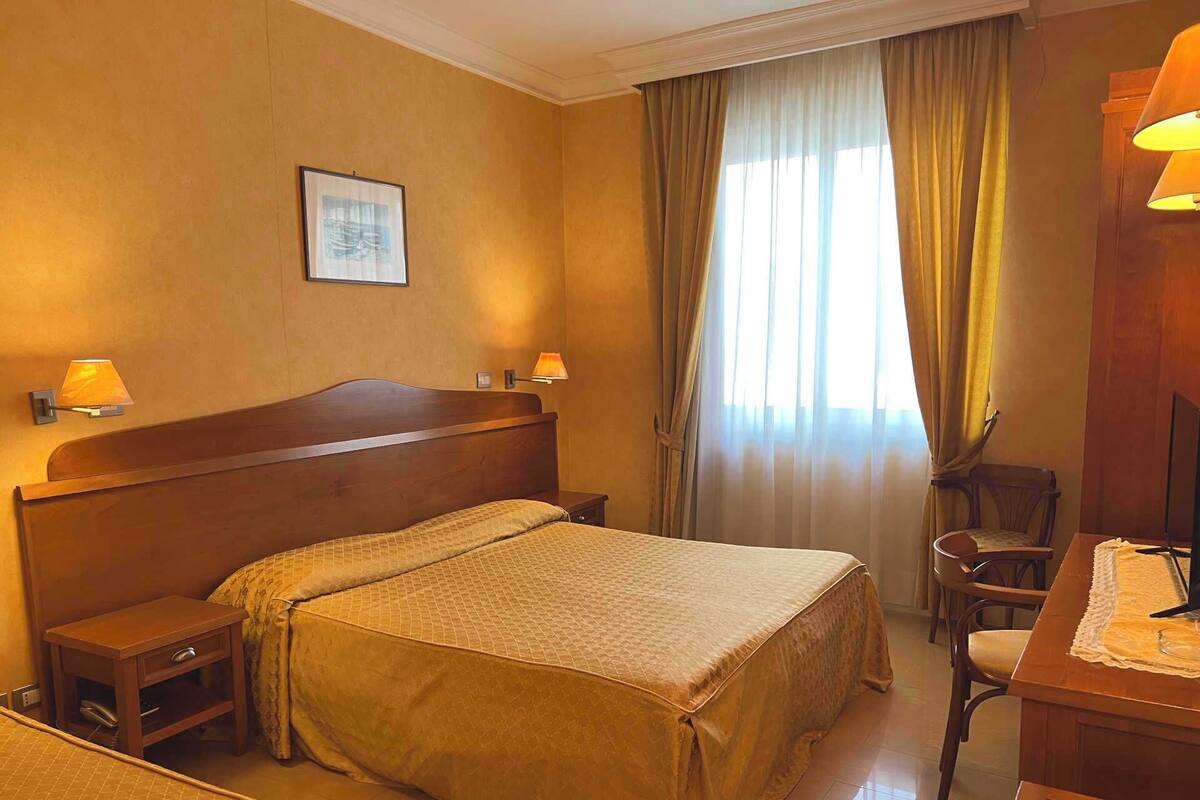 A hotel room with a bed, bedside tables, a desk with a chair, a lamp, and a window with curtains.