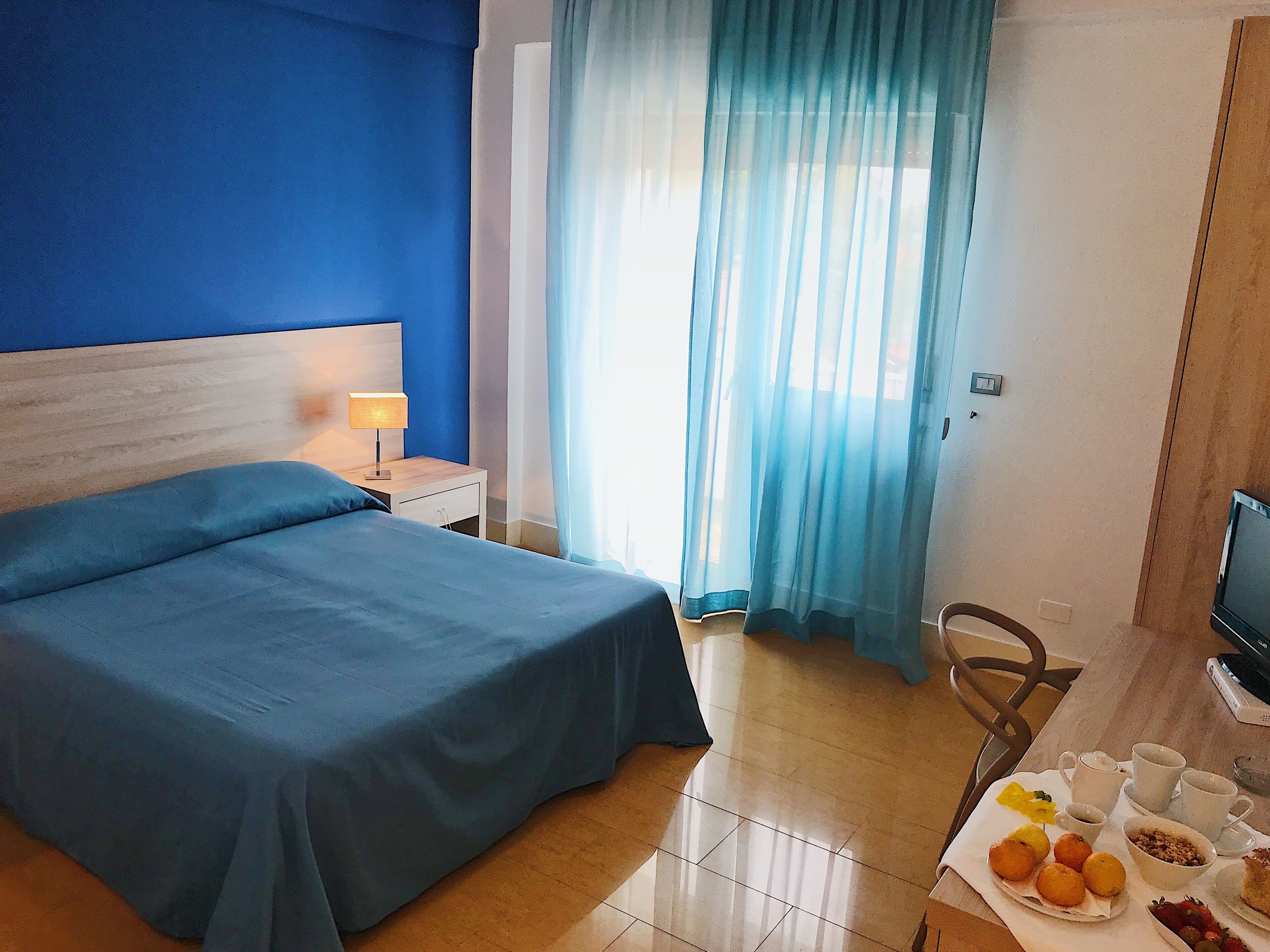 Une chambre d’hôtel comprenant un lit, une table de chevet avec une lampe, une télévision, une table à manger avec des articles pour le petit-déjeuner et une fenêtre avec des rideaux.