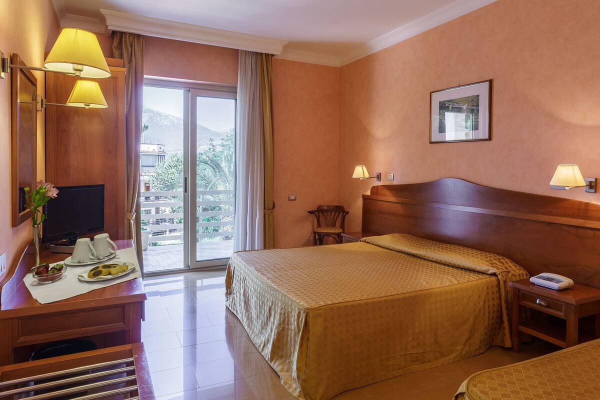 A hotel room with a bed, bedside table, lamp, and a desk with a TV and breakfast items.