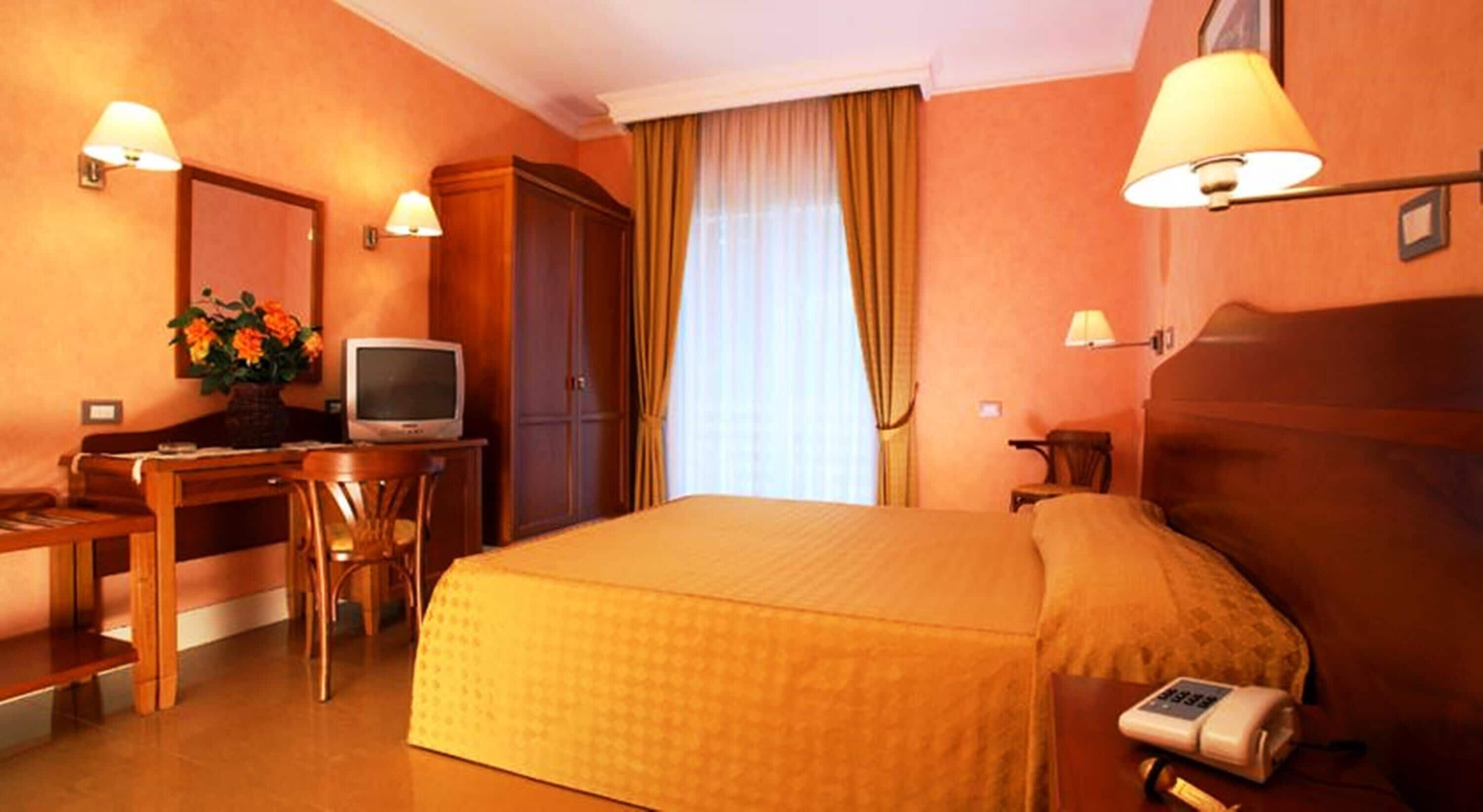 A hotel room with a bed, a desk with a television, a chair, a lamp, and a bedside table with a phone.