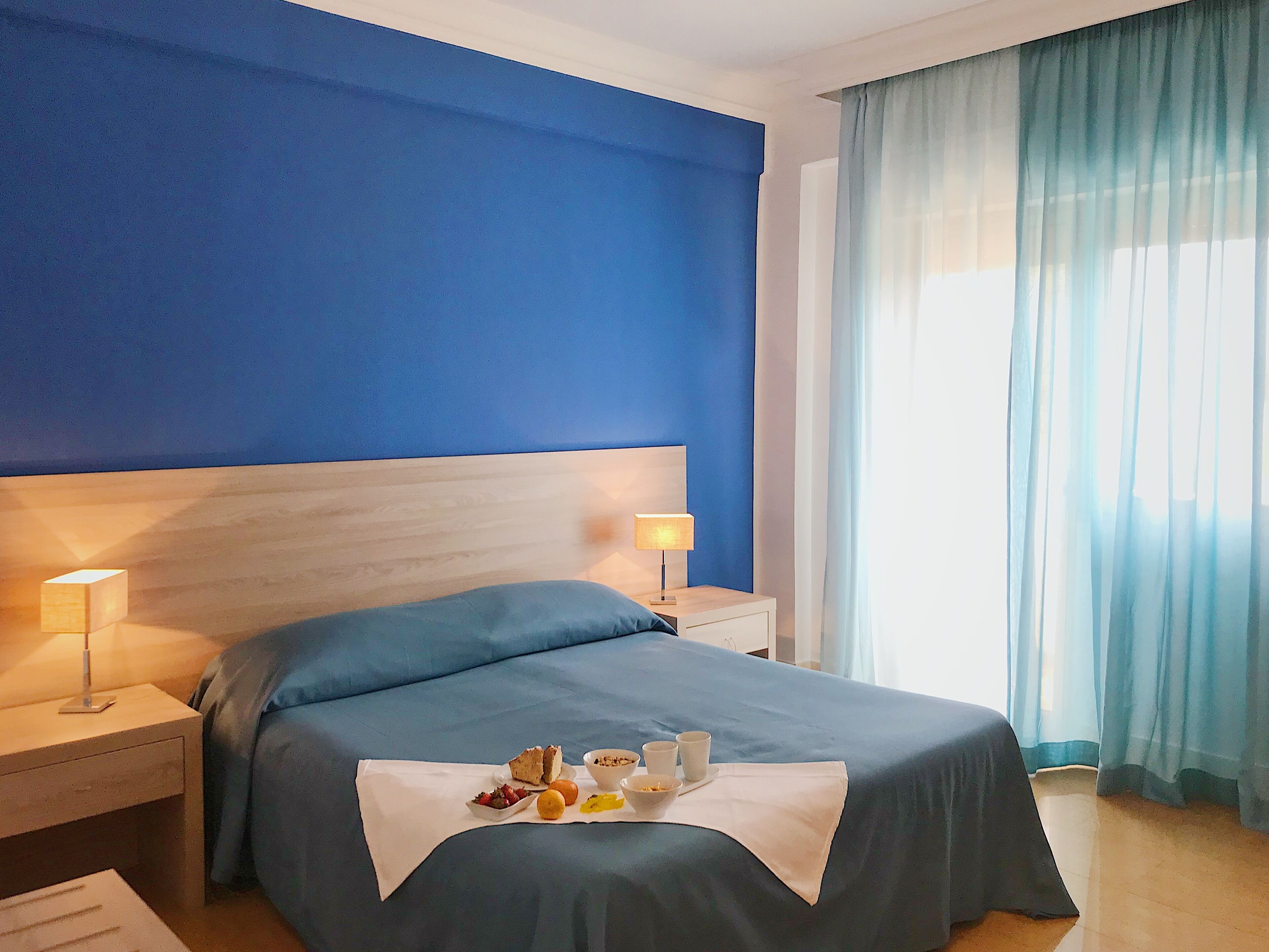 Ein Hotelzimmer mit einem Bett, Nachttischen, einer Lampe und einem Frühstückswagen mit Früchten und einer Schüssel.