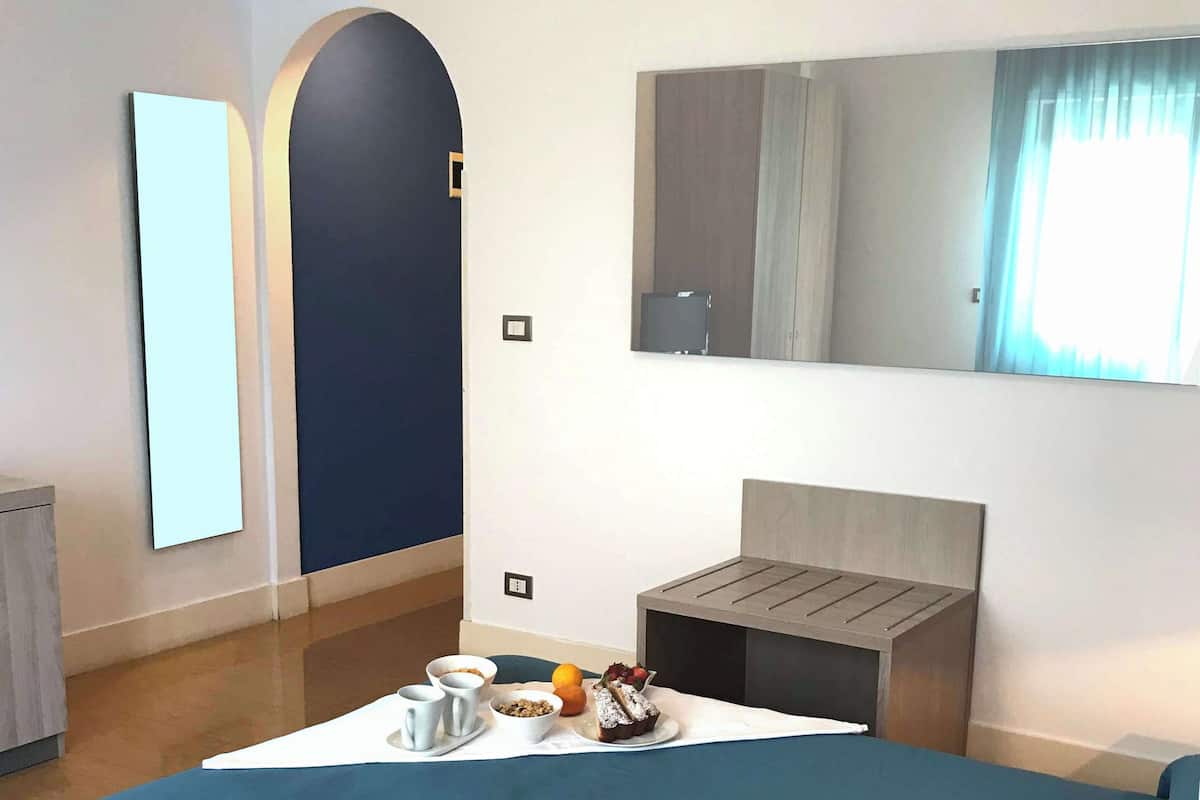 A modern hotel room with a bed, bedside table, and a small seating area. There is a large mirror on the wall and a window with curtains.