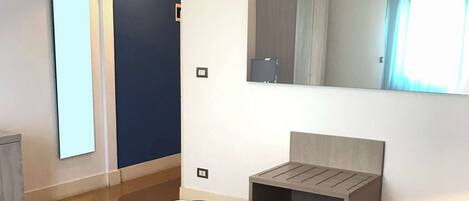 A modern hotel room with a bed, bedside table, and a small seating area. There is a large mirror on the wall and a window with curtains.