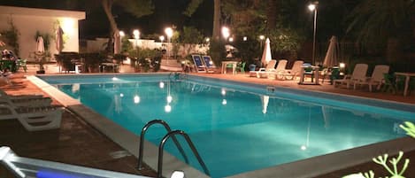 An outdoor swimming pool with lounge chairs and umbrellas, surrounded by trees and night lighting.