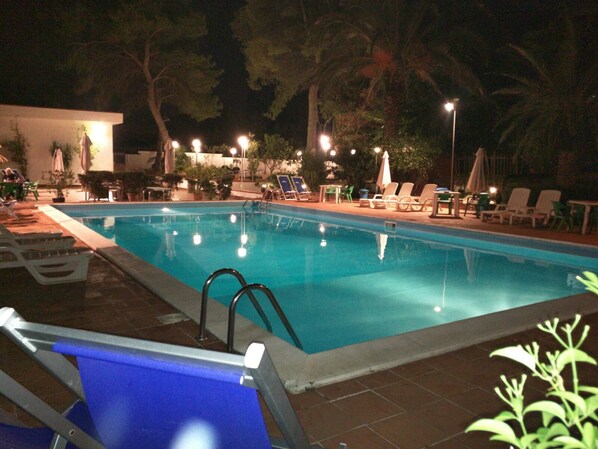 An outdoor swimming pool with lounge chairs and umbrellas, surrounded by trees and night lighting.