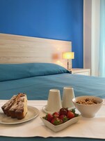 Un letto rifatto con cura, una testata blu, un comodino con una lampada e un vassoio colazione con torta, fragole e una ciotola di cereali.