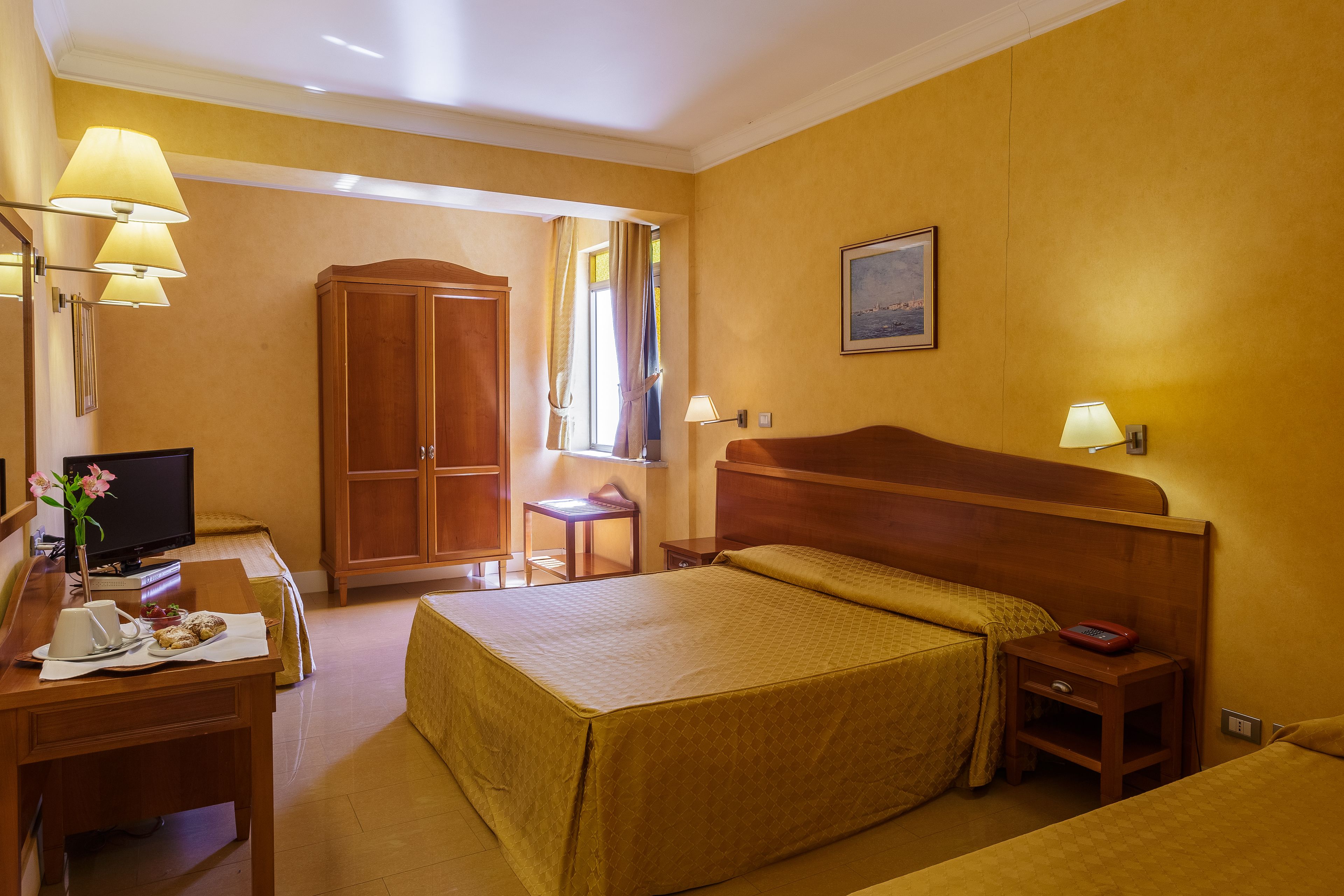 A hotel room with a bed, bedside table, lamp, desk with a television, and a wardrobe.