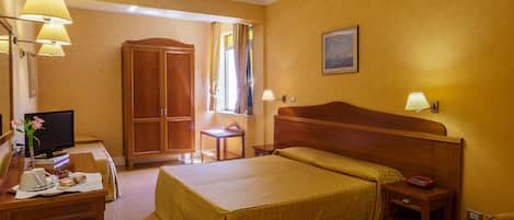 A hotel room with a bed, bedside table, lamp, desk with a television, and a wardrobe.