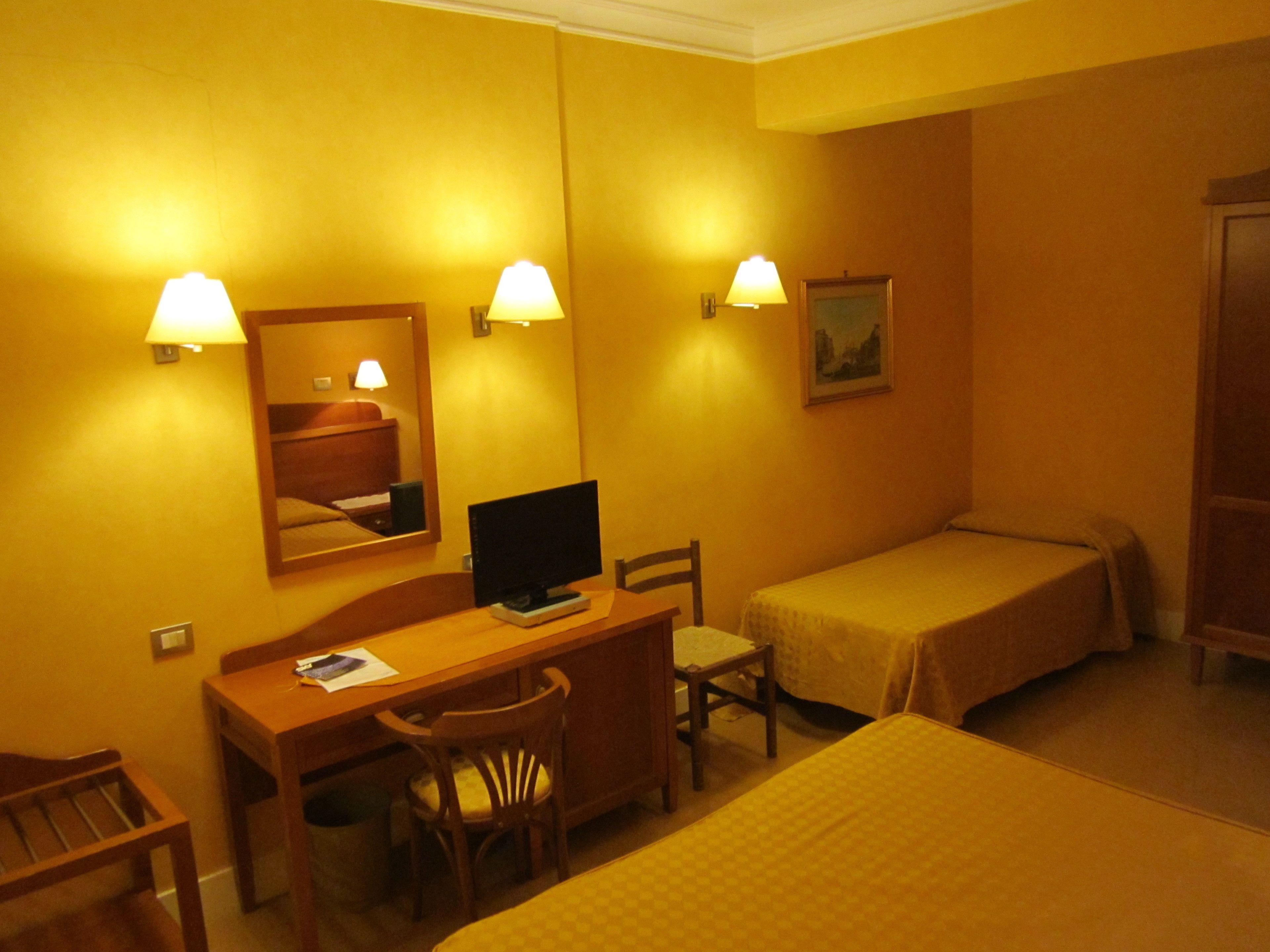 A hotel room with a desk, two single beds, a chair, a television, and wall-mounted lamps.