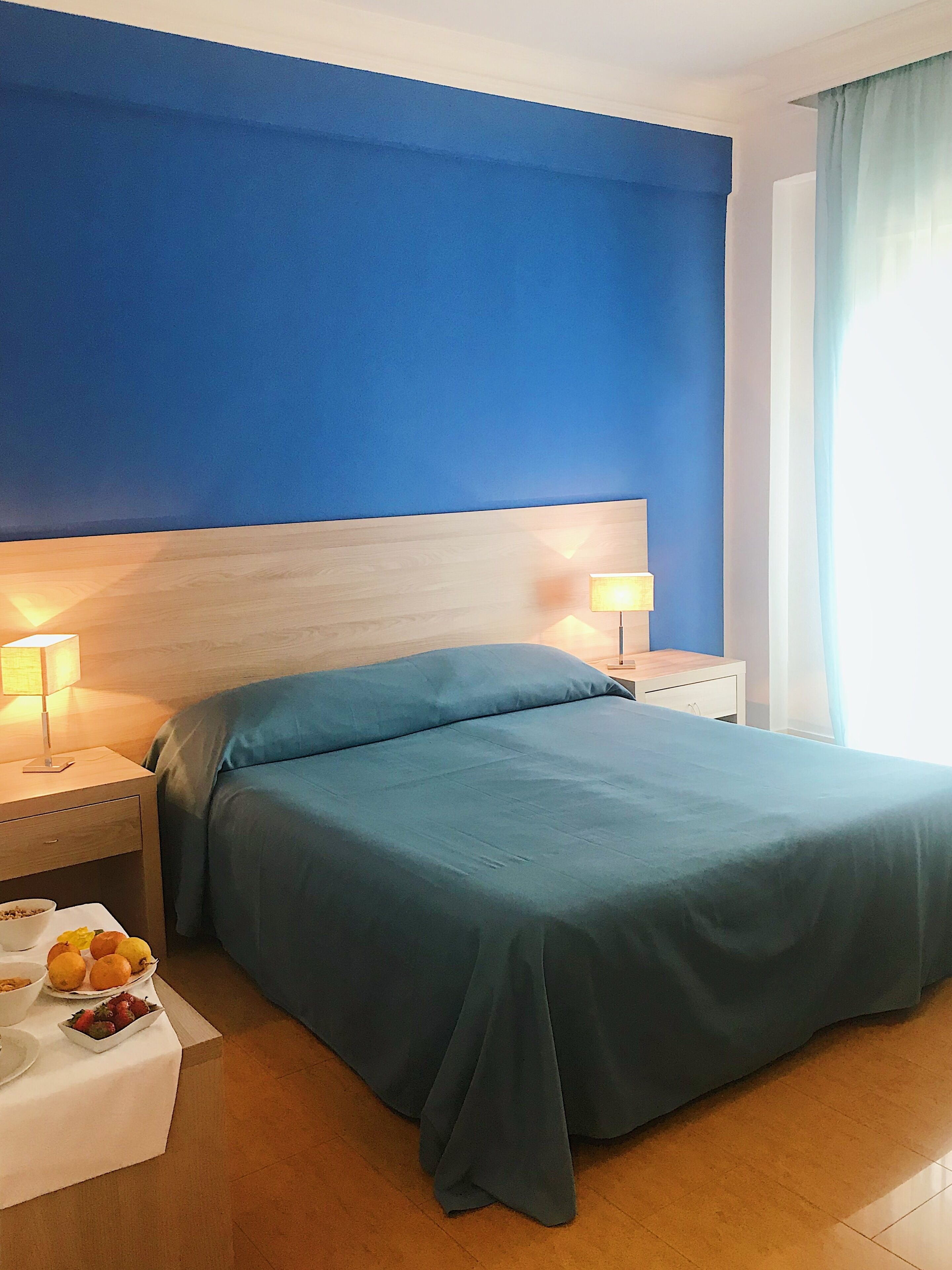 Une chambre d’hôtel avec un grand lit, deux tables de chevet avec des lampes, une petite table avec un panier de fruits et une fenêtre avec des rideaux.