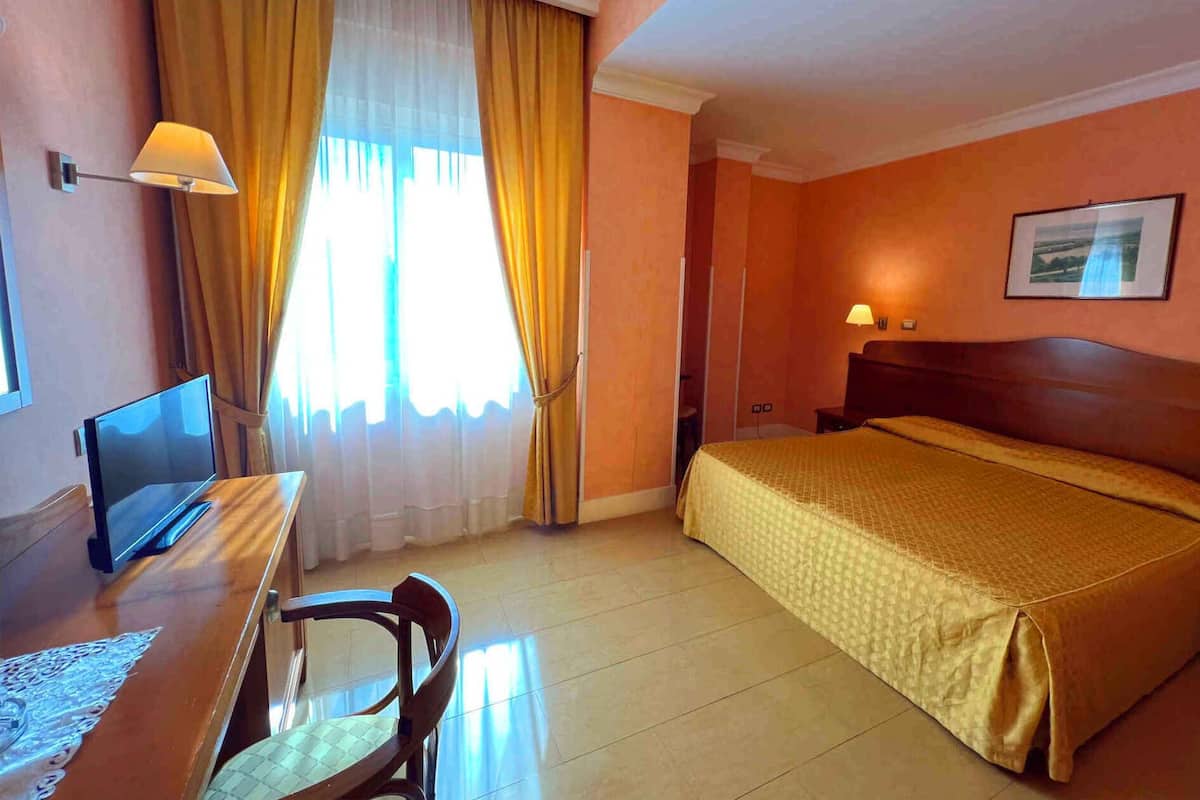 A hotel room with a bed, a desk with a computer, a chair, a window with curtains, and a wall-mounted lamp.