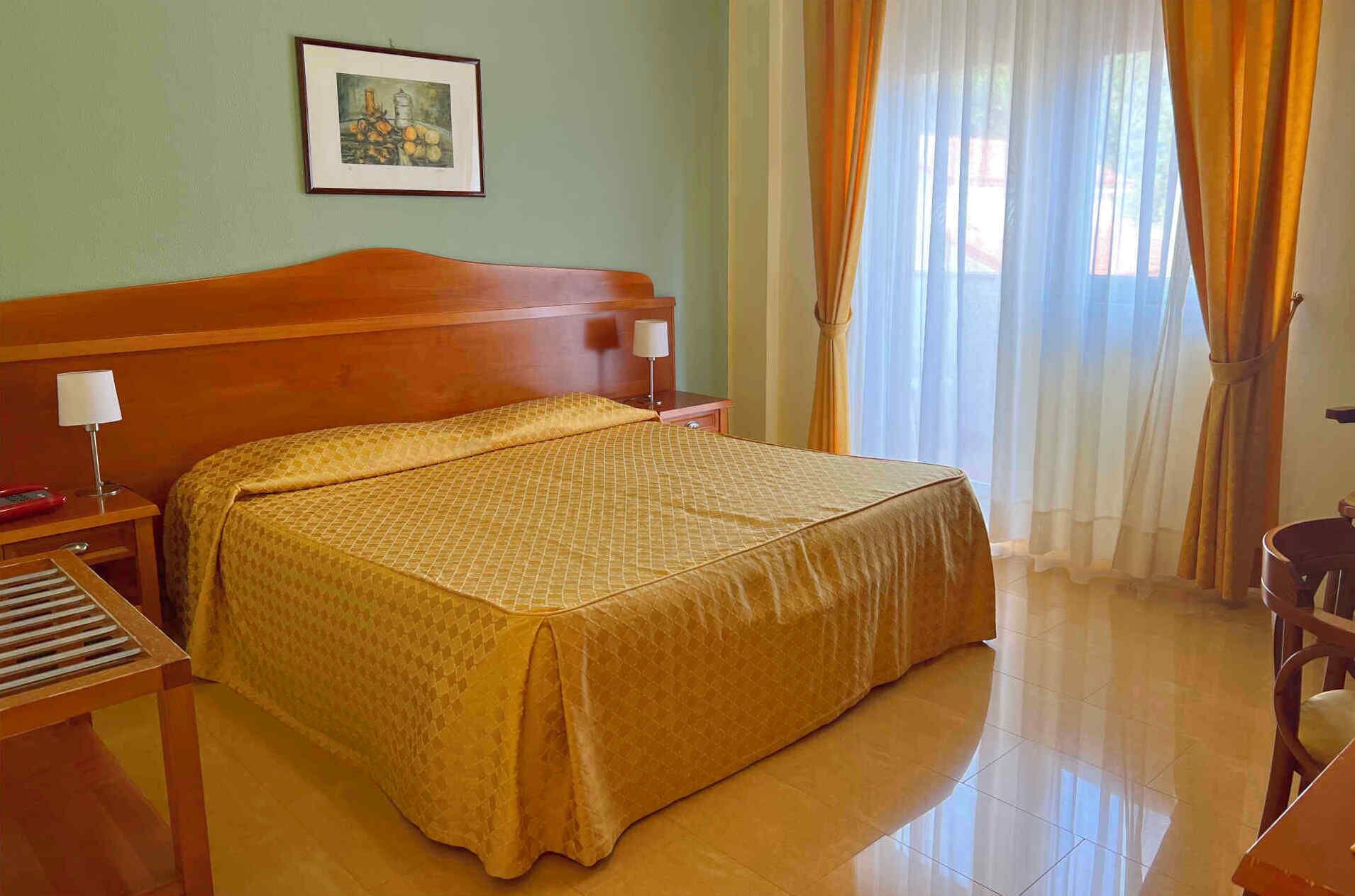 Una habitación de hotel con una cama grande, una mesita de noche con lámpara, un cuadro en la pared y una ventana con cortinas.