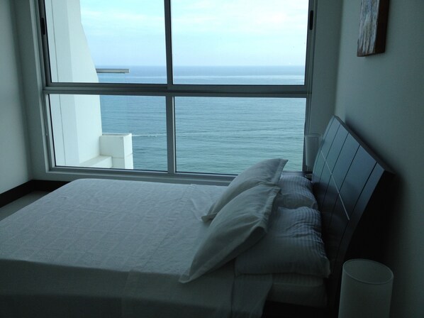 A bedroom with a large window overlooking the sea, a bed with white linens, and a bedside table.