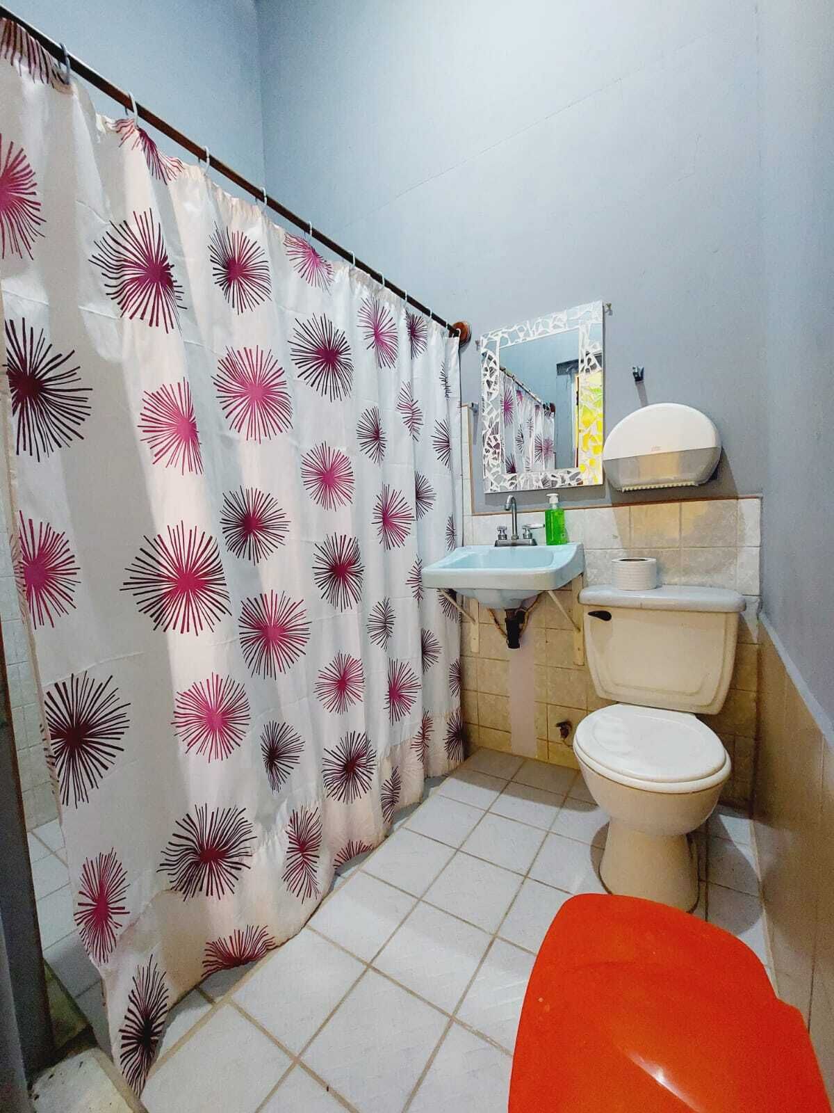 A bathroom with a shower curtain, sink, mirror, toilet, and a red chair.