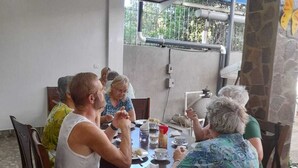 Un grupo de personas sentadas alrededor de una mesa en un área de comedor al aire libre techada, con pilares de piedra y vistas a la vegetación.