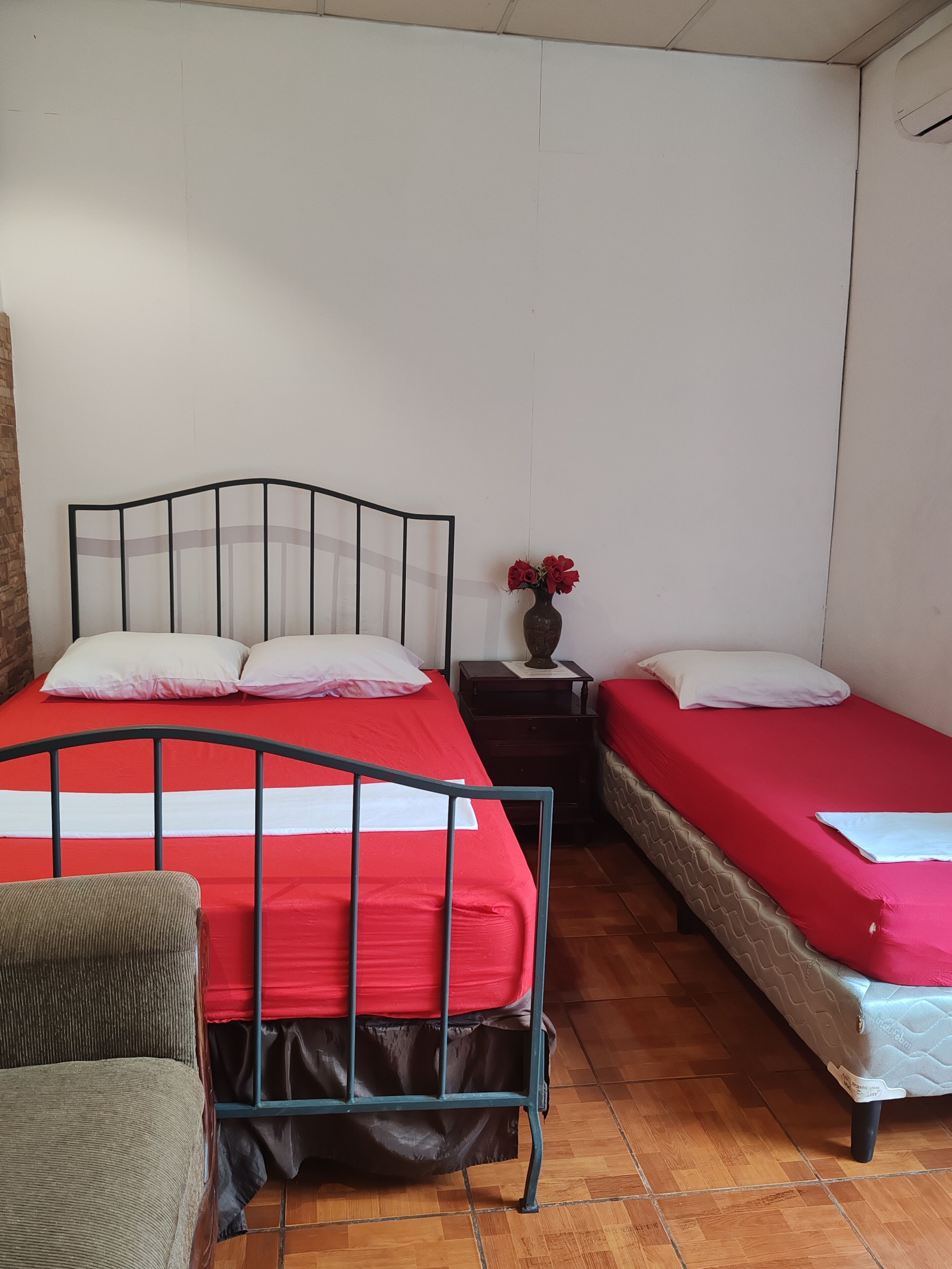 Una habitación con dos camas, una con colcha roja y la otra con colcha blanca. Hay una mesita con un florero, una silla y suelo de madera.
