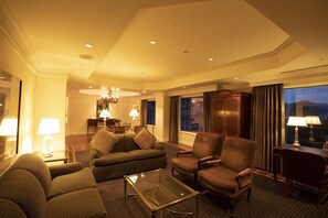 Ein Hotelzimmer mit Sofa, Sesseln, einem Glastisch, einer Chandelierleuchtre und Blick auf die nächtliche Stadt.