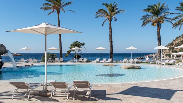 A pool area with lounge chairs and umbrellas, surrounded by palm trees and a view of the sea. - Falkensteiner Resort Capo Boi (Villasimius)
