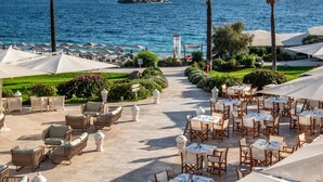 A seaside restaurant with outdoor seating, palm trees, and a view of the sea. - Falkensteiner Resort Capo Boi (Villasimius)