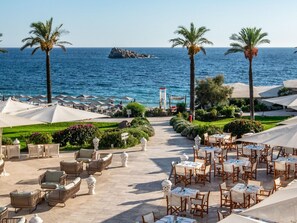 A seaside restaurant with outdoor seating, palm trees, and a view of the sea. - Falkensteiner Resort Capo Boi (Villasimius)