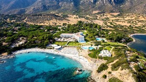 An aerial view of a coastal resort with a beach, mountain backdrop, and a large resort building. - Falkensteiner Resort Capo Boi (Villasimius)
