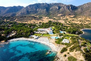 An aerial view of a coastal resort with a beach, mountain backdrop, and a large resort building. - Falkensteiner Resort Capo Boi (Villasimius)