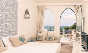 A bedroom with a large bed, a view of the sea through a window, and a balcony with a railing. - Falkensteiner Resort Capo Boi (Villasimius)