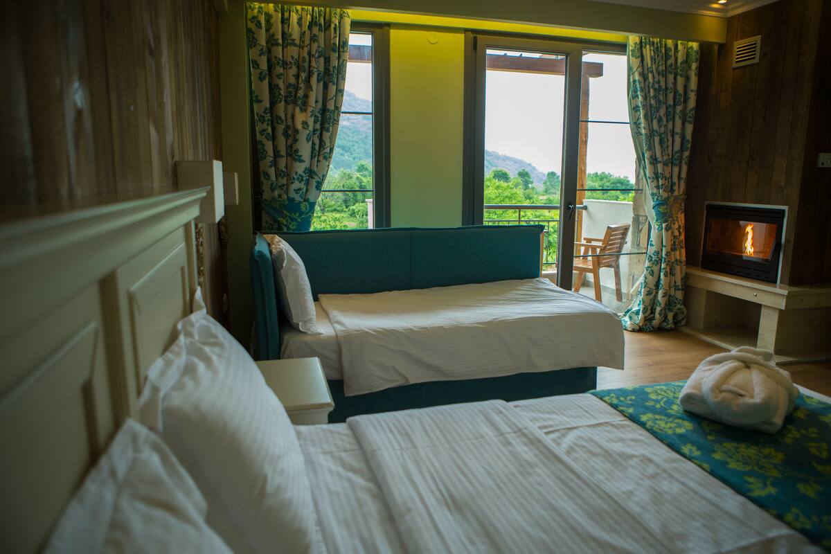 A hotel room with two beds, a sofa, a fireplace, and a balcony with a view of greenery.