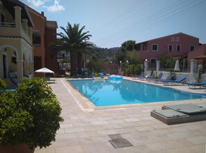A swimming pool with lounge chairs and inflatable floats, surrounded by buildings and palm trees. - Katerina Pool Apartments (Corfu)