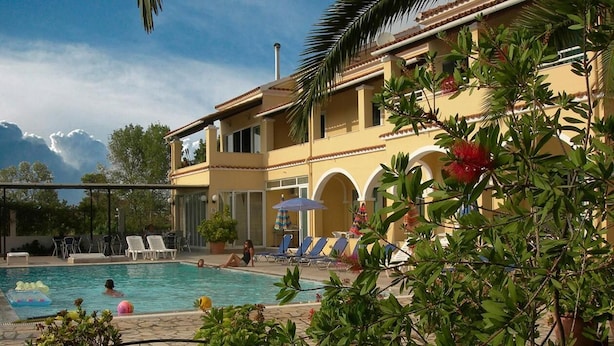 Zona de piscina con sillones de descanso y sombrillas, rodeada de vegetación y un edificio con arcos.