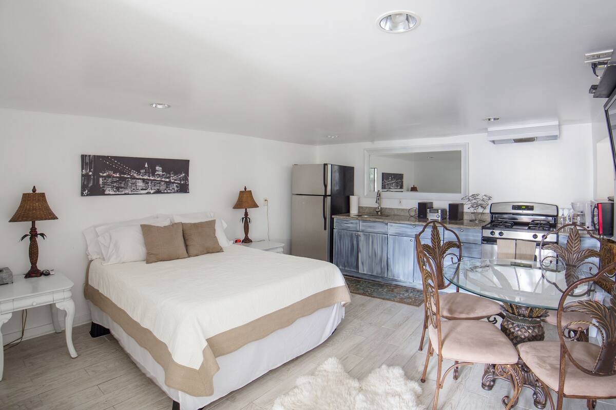 A hotel room with a bed, a desk, a dining table with chairs, a kitchen area, and a bathroom.