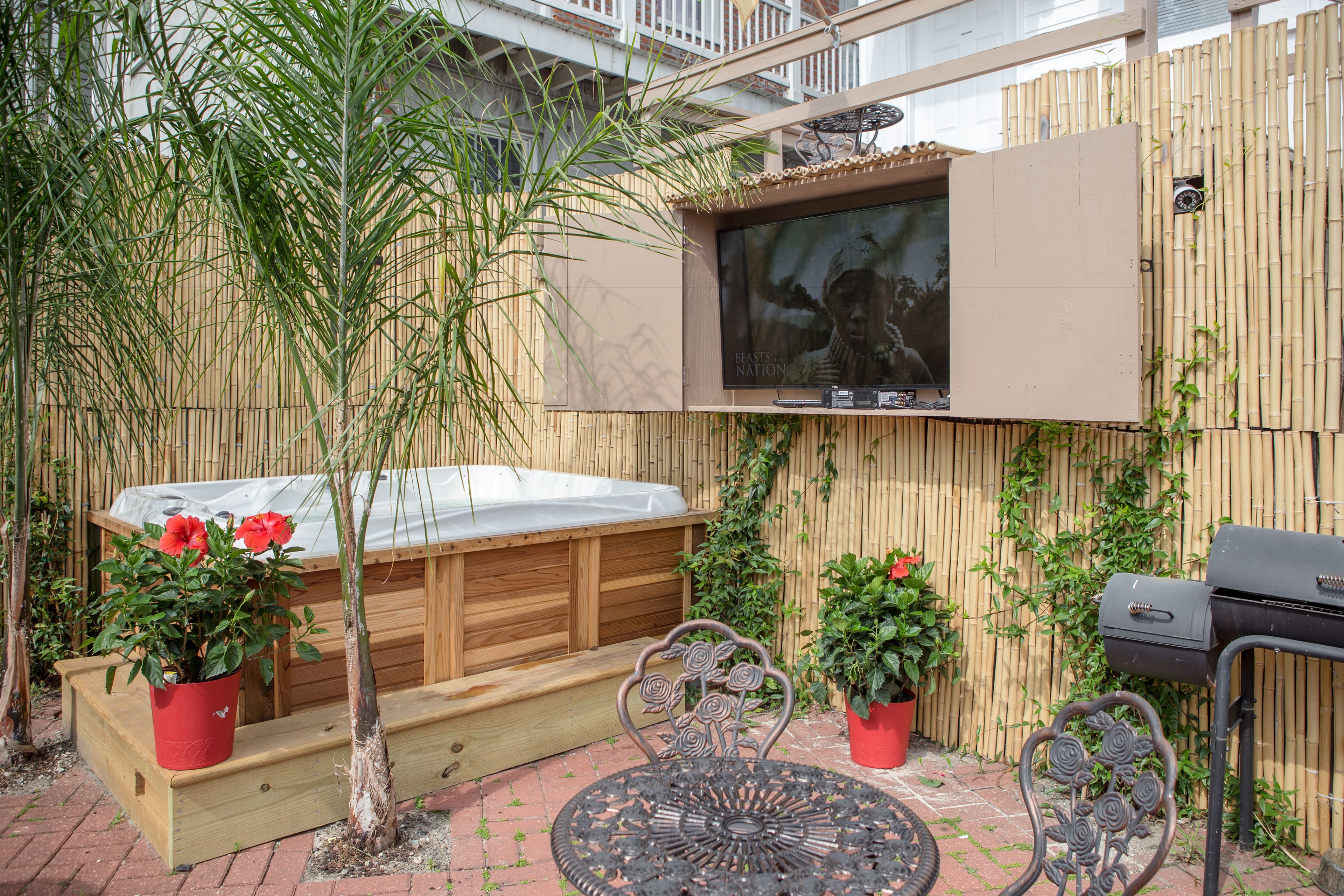 A patio area with a hot tub, wooden deck, outdoor seating, and a TV mounted on a bamboo wall.