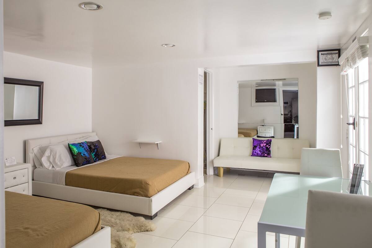 A modern bedroom with two beds, a sofa, a glass table, and a kitchen area visible through an open door.