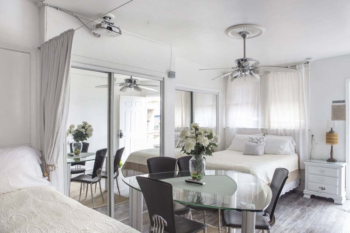 A hotel room with a glass dining table, two black chairs, a bed with white bedding, a ceiling fan, and a nightstand with a lamp.