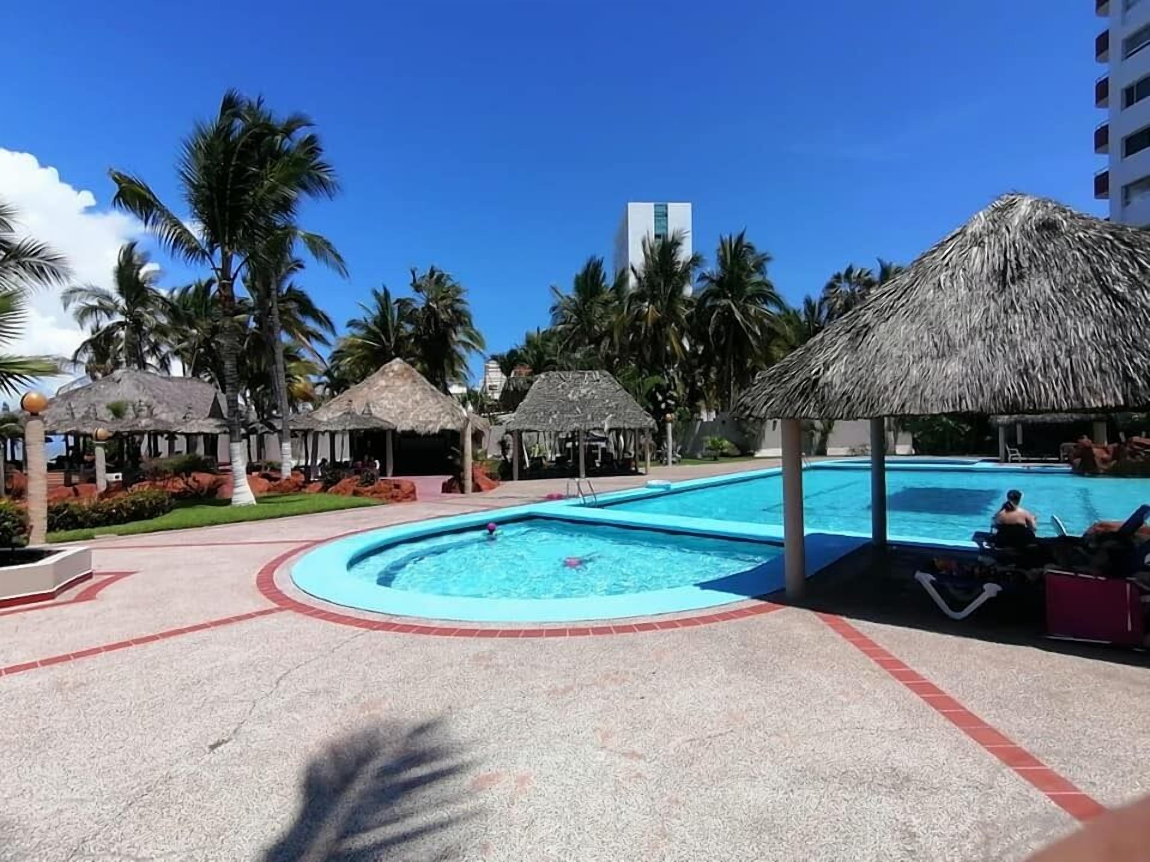 A resort pool area with a circular pool, thatched-roof structures, palm trees, and a tall building in the background.